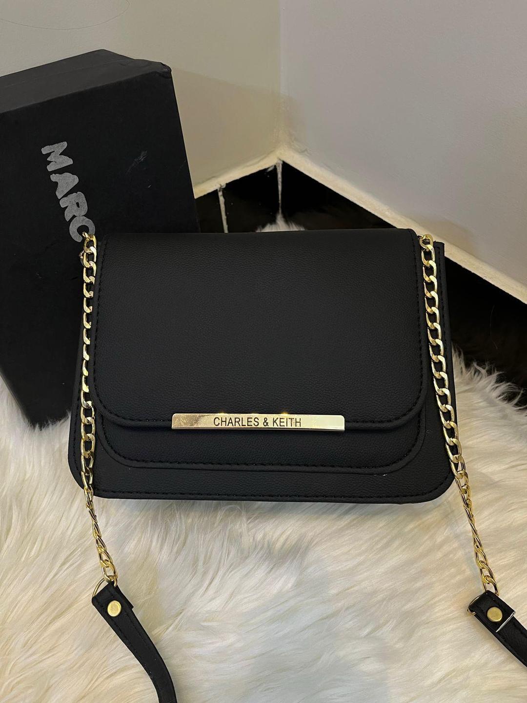 Black handbag with gold chain strap on a white fur surface, with 'Marc' and 'Charles & Keith' branding visible.