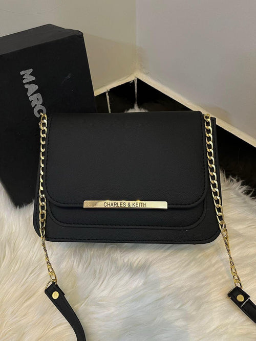 Black handbag with gold chain strap on a white fur surface, with 'Marc' and 'Charles & Keith' branding visible.