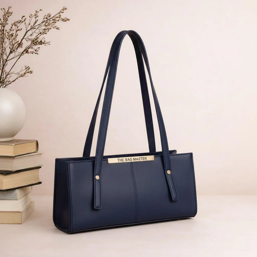 Navy blue handbag with gold accents on a beige surface with books and a decorative vase.